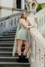Plus size model in Candice sage green mini dress leaning on stairs, showcasing the dress's flattering fit and long chiffon scarf for semi-formal occasions.