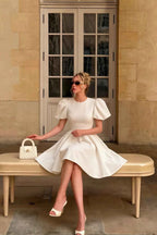 Customer in LVLVS Celia white stretch crepe crew neck puff sleeve mini dress sitting on bench, elegant day party look with white handbag and pointed toe heels