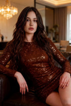 Model in LVLVS Charlotte Chocolate Sequin Dress sitting on leather sofa, casual luxe style for homecoming or brunch, long sleeves and round neck, rich sequin fabric catching light in cozy lounge setting.
