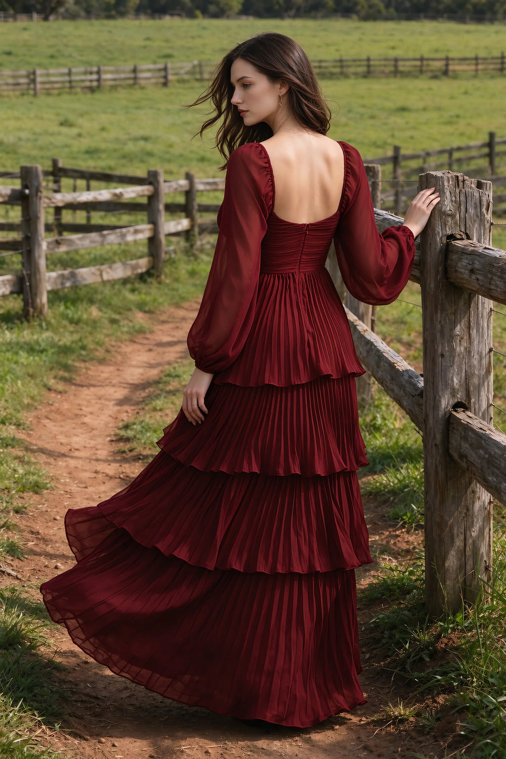 Outdoor rear view by a countryside fence of Sienna burgundy tiered pleated maxi dress with a long flowing skirt