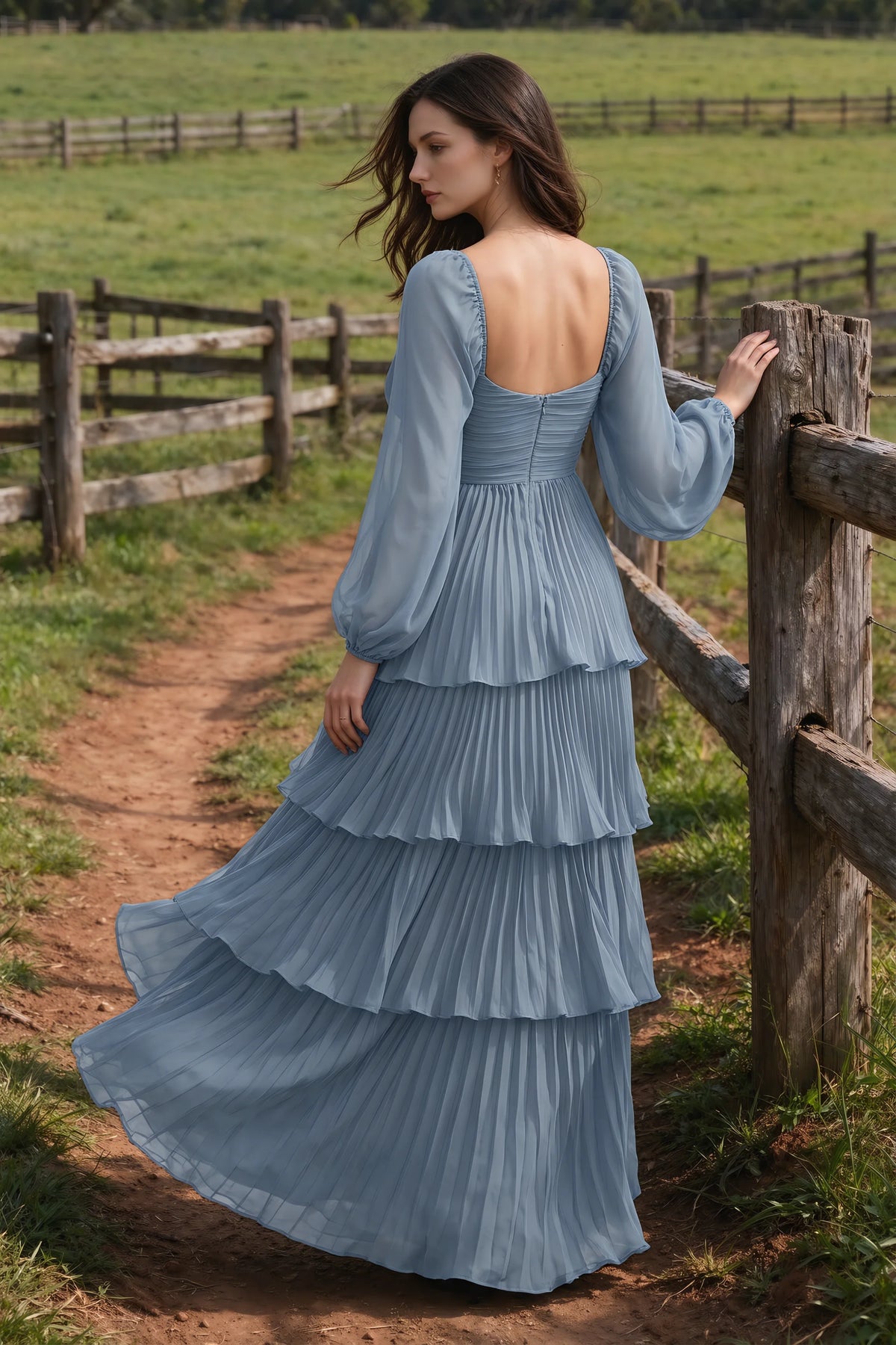 Outdoor rear view by a countryside fence of Sienna dusty blue tiered pleated maxi dress with a long flowing skirt