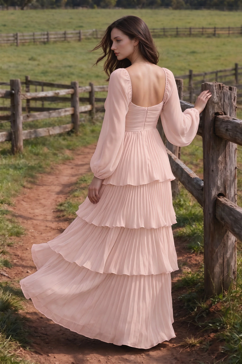 Outdoor rear view by a countryside fence of Sienna haze pink tiered pleated maxi dress with a long flowing skirt