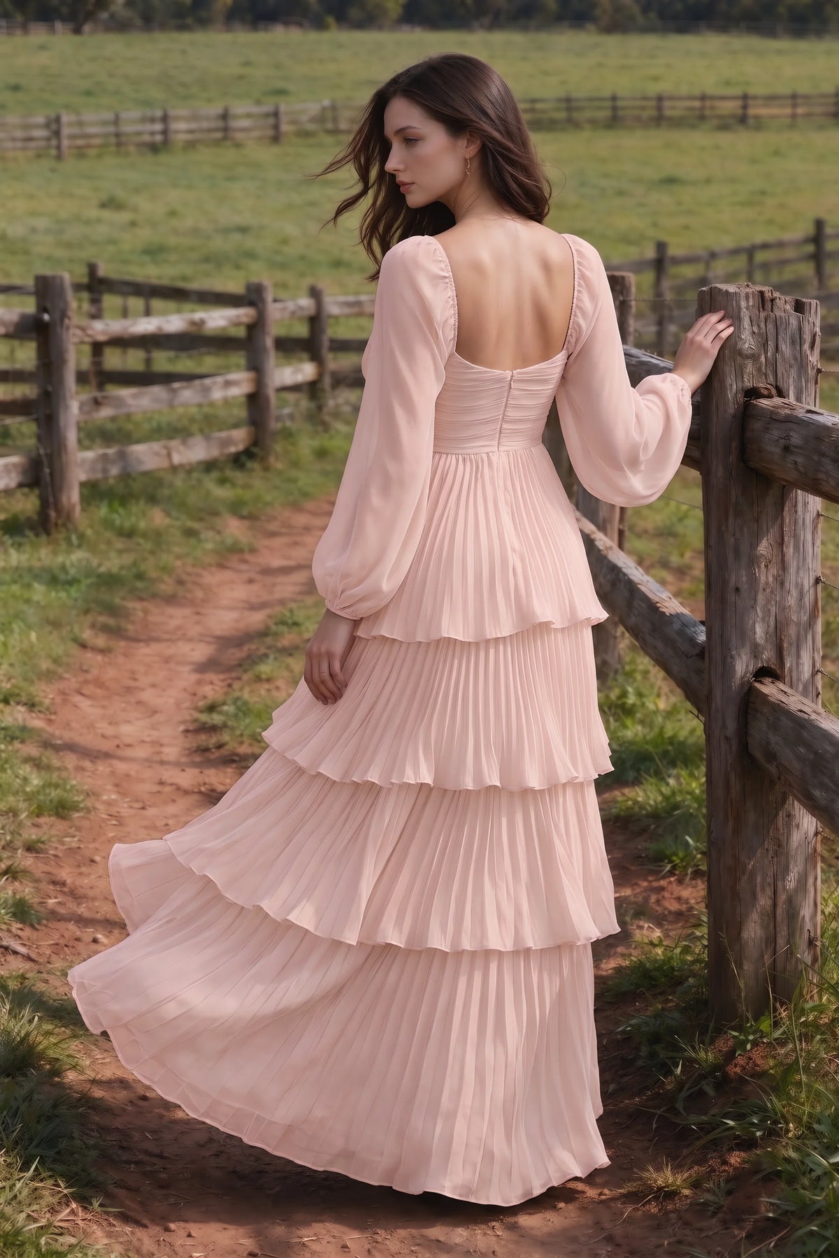Outdoor rear view by a countryside fence of Sienna haze pink tiered pleated maxi dress with a long flowing skirt
