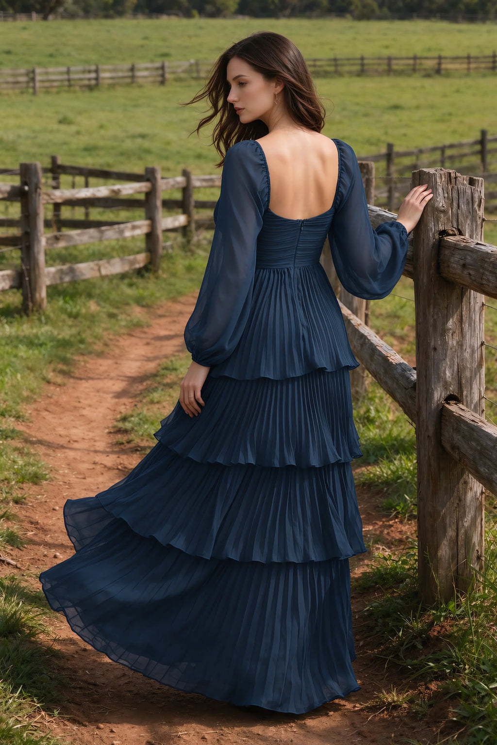 Outdoor rear view by a countryside fence of Sienna navy tiered pleated maxi dress with a long flowing skirt