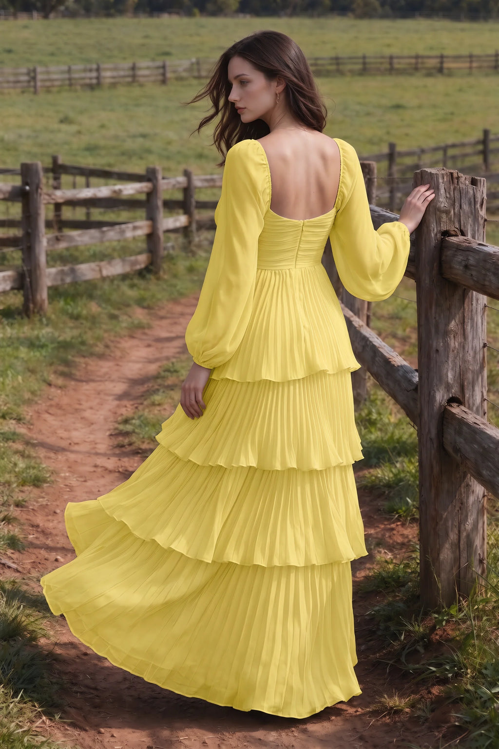 Outdoor rear view by a countryside fence of Sienna pastel yellow tiered pleated maxi dress with a long flowing skirt