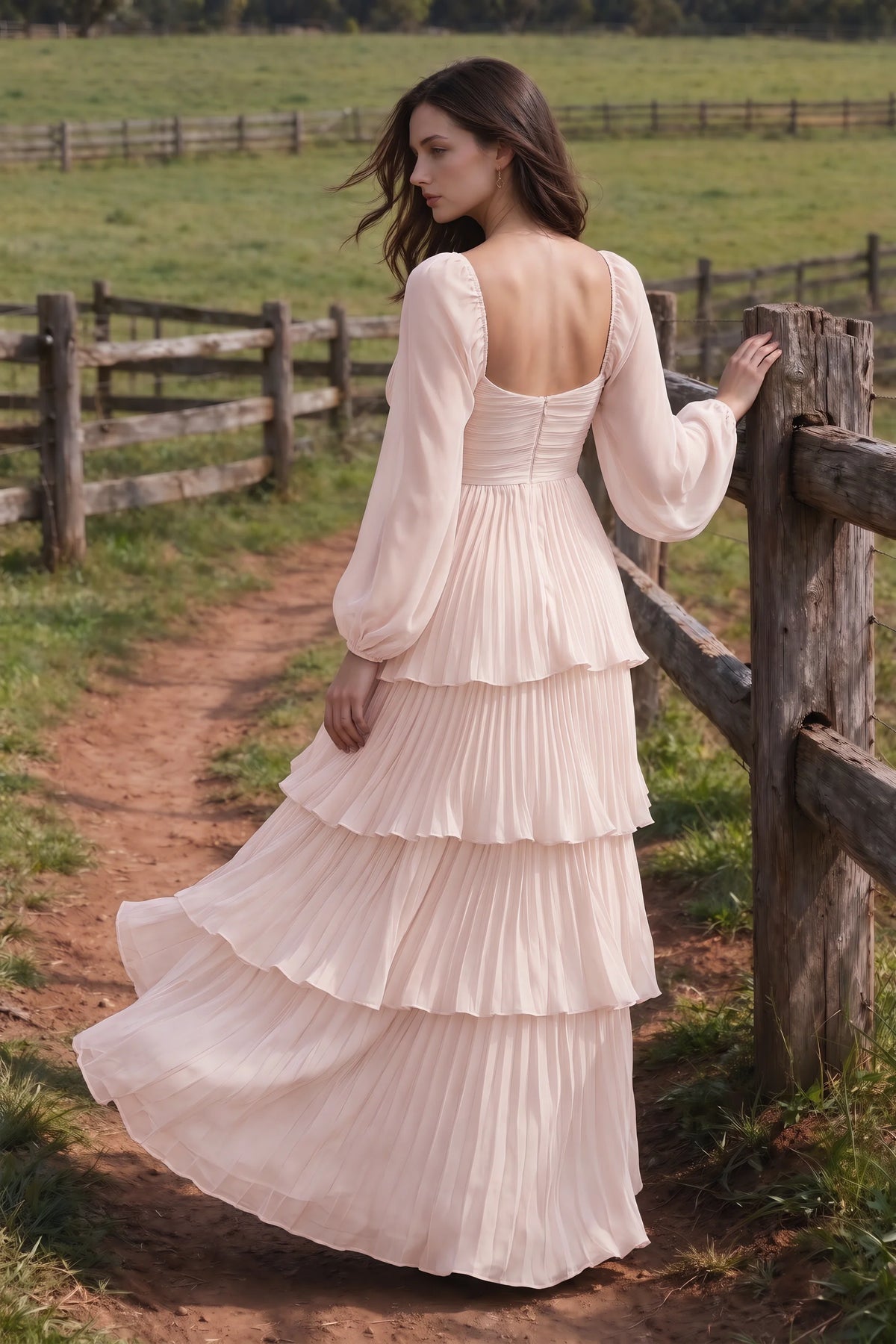 Outdoor rear view by a countryside fence of Sienna powder pink tiered pleated maxi dress with a long flowing skirt