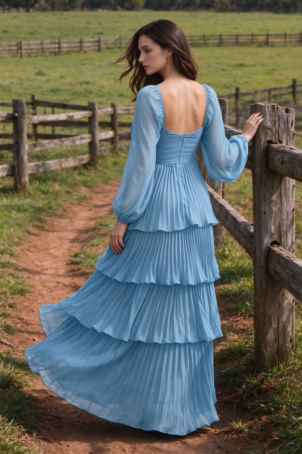 Outdoor rear view by a countryside fence of Sienna steel blue tiered pleated maxi dress with a long flowing skirt